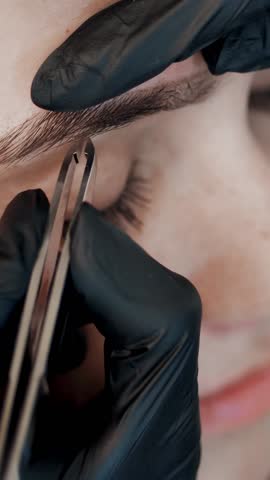 Hands of the cosmetician in black rubber gloves hold tweezers and pull out eyebrows. Master corrects the shape of the eyebrows. Beauty saloon of brow artist.