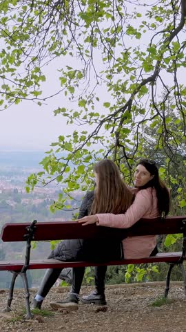 Mother and daughter, a woman with a teenager girl are sitting on a bench in the park, talking, relaxing. spring. flowering trees with fresh green leaves. beautiful view, landscape.