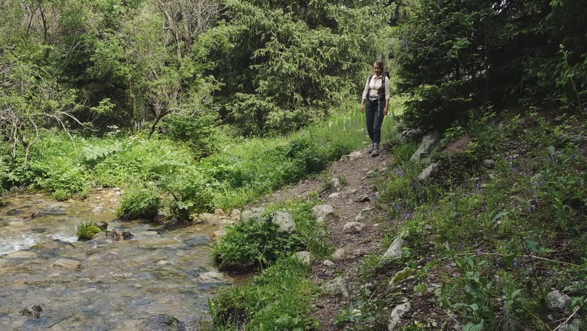 A tired backpacker and her dog approach a mountain river during a hike on a hot sunny summer day. The woman washes her face and the dog drinks water. National Park in Almaty, Kazakhstan.
