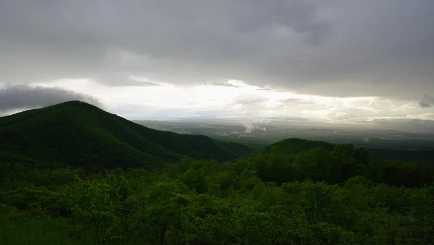 Panoramic view of rolling green mountains and valleys at sunset in Shenandoah National Park, Virginia, with dramatic storm clouds and soft golden light breaking through the sky.