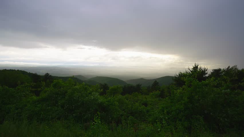 Panoramic view of rolling green mountains and valleys at sunset in Shenandoah National Park, Virginia, with dramatic storm clouds and soft golden light breaking through the sky.