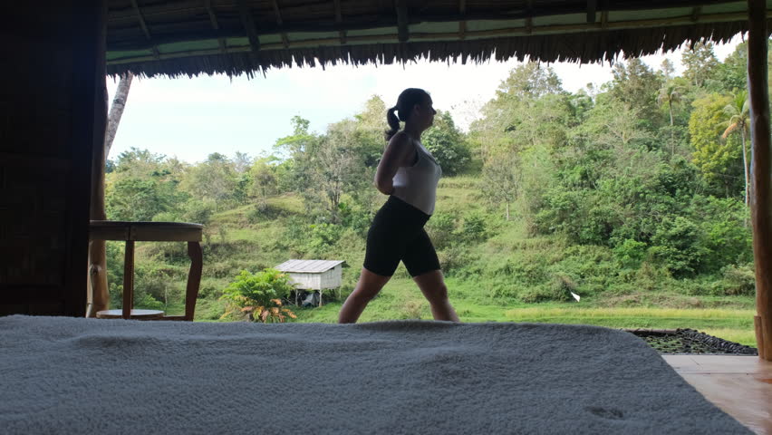 Young woman practices yoga on a serene terrace of tropical hut, connecting with calmness of nature amidst rice fields and vibrant greenery. Active lifestyle and wellness concept