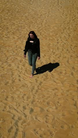 Woman walking barefoot on sandy beach or desert, vertical orientation. Female moving calmly across golden sand in sunlight. Lady strolling alone through arid terrain or coastal area
