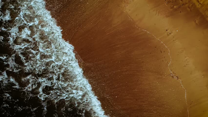 Foamy wave covering smooth golden shore. Sea water sliding across bright wet sand. Ocean surf reaching soft coastline in top view. Curling tide creating white border on beach surface