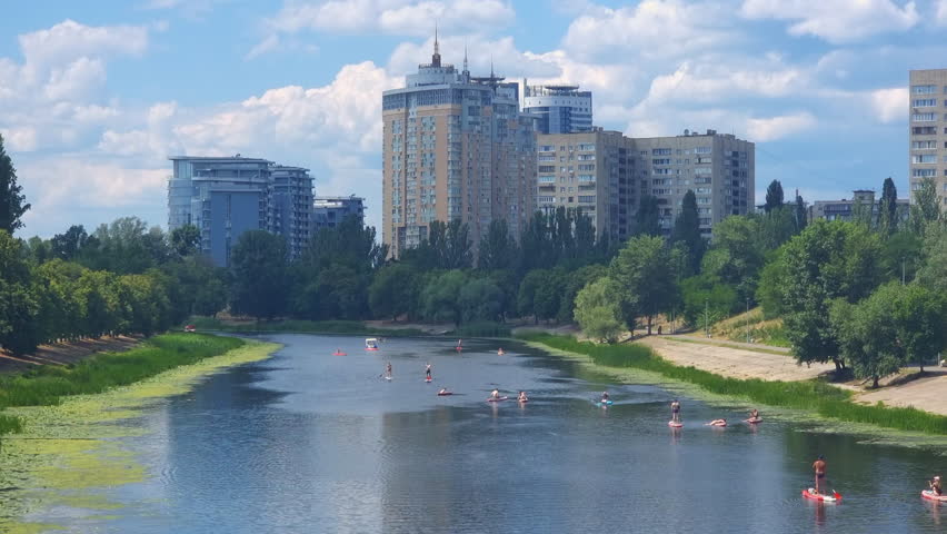 Residential high-rise buildings lining Dnipro riverbank, showcasing urban recreation with people paddling on stand up paddleboards during sunny summer day in Kyiv, Ukraine
