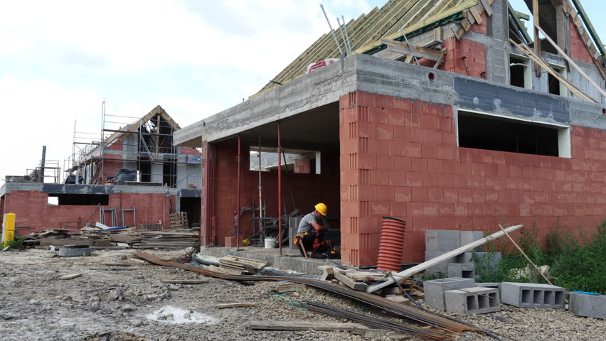 A construction worker carefully places roof trusses on a new building while standing on a scaffold. The clear blue sky provides excellent working conditions.
