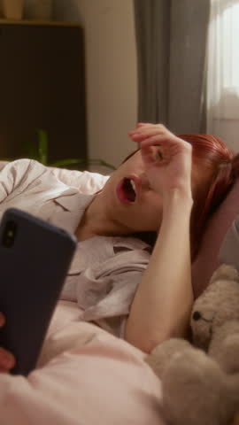 Vertical shot of bored young red-haired Caucasian girl yawning while chilling in bed with her smartphone