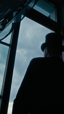 Vertical back-view shot of vintage-styled man in formal attire adjusting his cylinder hat with gloved hands as he standing by window, gazing thoughtfully at clear blue sky beyond