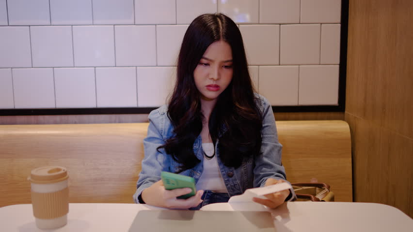 In a restaurant, young asian woman sits alone, visibly stressed as she reveals her bills and calculates expenses on her phone, striving to manage her finances effectively.