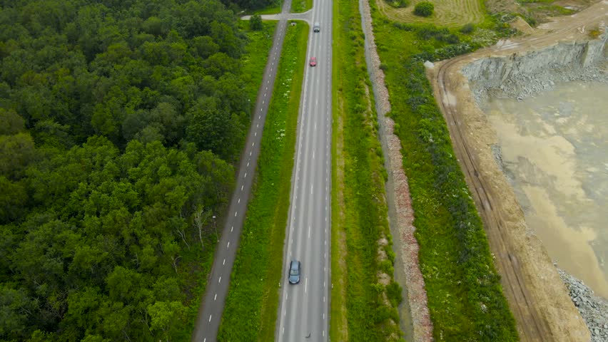 Aerial drone footage descending and flying lower above a rural countryside highway where cars and vehicles are driving at a fast speed during summer day. A large limestone quarry next to the road.