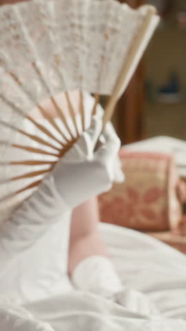 Vertical of aristocratic lady waving lace fan while seated on plush sofa surrounded by opulent furnishings, embodying grace, charm, and timeless luxury