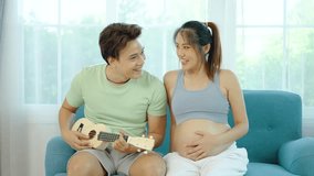Beautiful pregnant asian woman and young man are smiling and playing Ukulele or acoustic guitar on sofa, Husband playing Ukulele for pregnant woman and baby in the womb, concept relax day and holiday - Powered by Shutterstock - Get 15% off with code: PIKWIZARD15