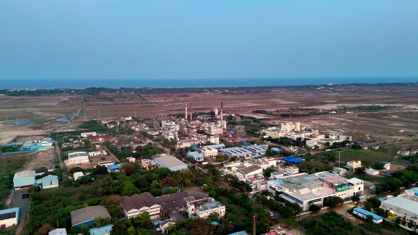 Drone footage capturing a blend of industrial and urban growth in Chennai, Tamil Nadu. Factories and buildings are scattered across the landscape, with the sea visible on the distant horizon.
