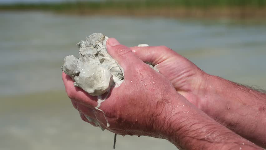 Close-up, natural blue clay in male hands. Clay from the lake bottom. Blurred water background on a sunny day. Uses, significance, and folk medicine.