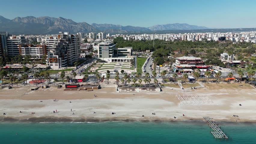 Drone flyover along Konyaalti Beach. Mountains, city skyline, pebble shore, and sea in one continuous coastal scene in Antalya.