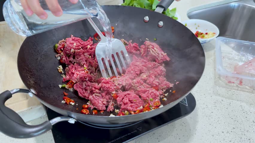 Raw minced meat is stir-fried with chopped chili peppers and vegetables in a wok, under bright kitchen lighting, with visible steam and utensil movement