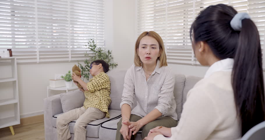 Asian single mother expresses frustration while sitting on sofa as female psychologist calmly listens and explains during therapy session about managing child adhd behavior and emotional stress