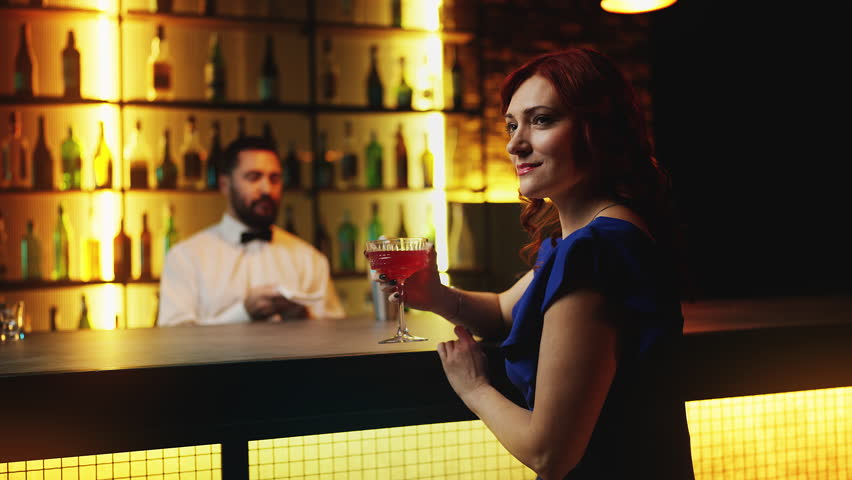 Playful redhead woman sipping cocktail at bar counter, looking around for a date
