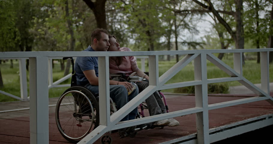 A tender moment shared between an inclusive couple in wheelchairs, finding comfort and love on a bridge in a serene green park. Their connection highlights mutual support and affection in nature.