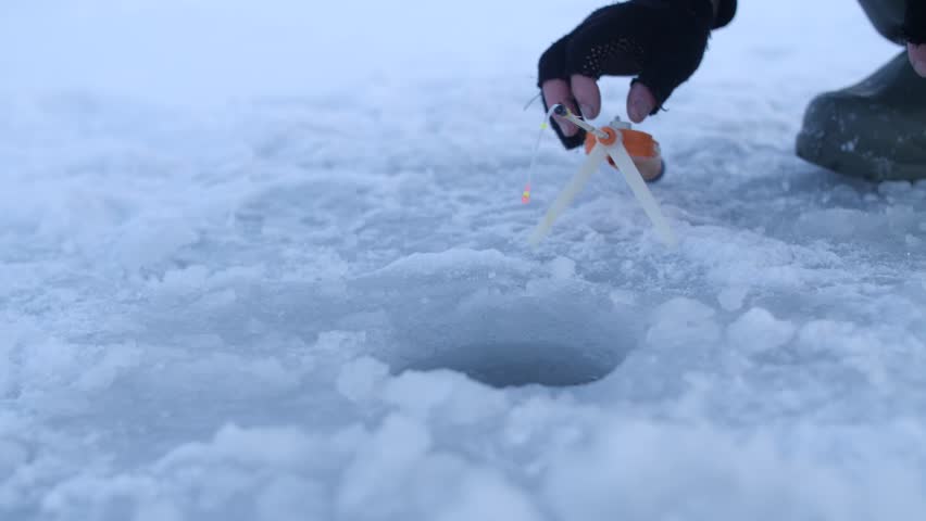 Fisherman carefully sets his fishing rod on frozen surface of lake near hole, preparing for ice fishing. Tranquil winter snowy landscape around. Hobby sports outdoors activity concept. Catching fish.