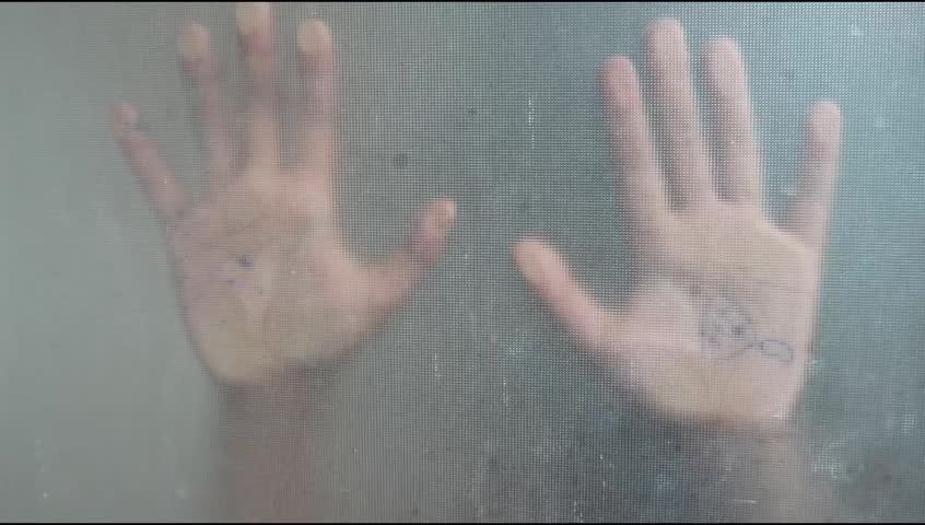 Two palms are pressed against a frosted, glass-like surface, creating a faint silhouette with a dramatic effect. One hand appears to have a pen or scribble on its palm, creating a mysterious and artis