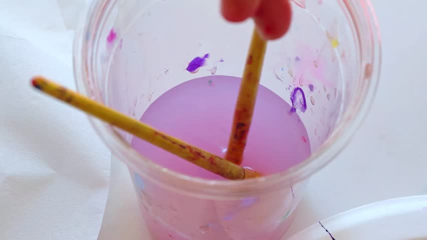 Cleaning paint brush in water in transparent cup. A child washes a paint brush in glass of water after painting. A view from above. 