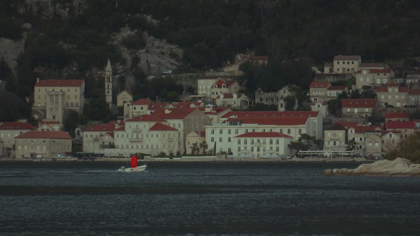 A lone boater glides through calm waters by a scenic village at dusk, highlighting beautiful architecture of Perast city in Montenegro