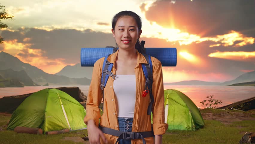 Asian Female Hiker With Mountaineering Backpack Smiling And Pointing To Side While Standing While Tent Camp Lakeside