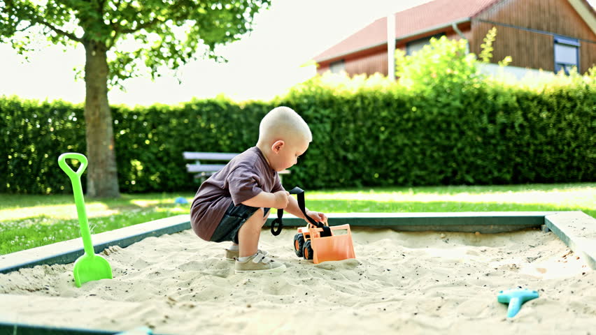Toddler in a t-shirt and shorts. Boy plays with a plastic car in the sand. Little 2 year old boy plays in the sandbox on a playground outdoors. Toddler plays with a sand shovel.