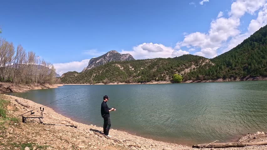A fisherman in black attire casts a fishing line into a tranquil mountain lake surrounded by lush pine forests and rugged hills under a partly cloudy sky.