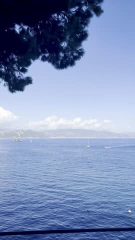 An image of a vibrant blue sea under a clear sky with white clouds, distant boats sailing, and far-off mountains completing the serene and picturesque coastal landscape.
