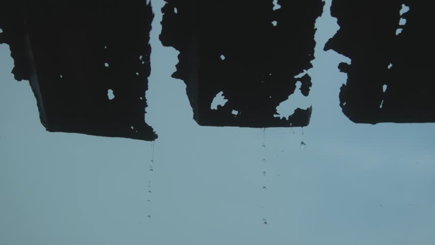 Water drops fall slowly from jagged, torn corrugated roofing panels silhouetted against a pale blue sky, capturing a minimal and somber moment of decay, texture, and quiet rhythm in nature.
