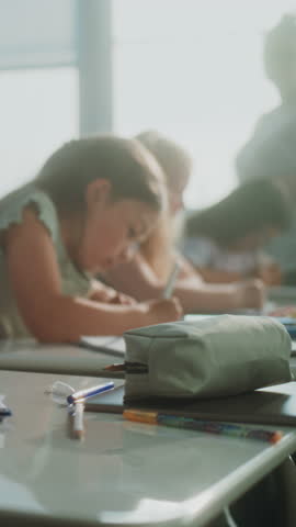 Primary School Children Writing School Test, Doing Tasks in Notebooks. Female Teacher Walking Between Desks, Controlling Group of Diverse Kids During Exam Process in Modern Classroom. Vertical Shot.