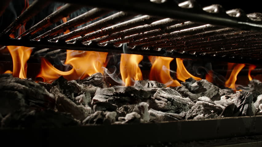 Spiced chicken fillet drops onto a hot grill with fire and smoke. Seasoning scatters as meat hits the grates. Camera moves up from burning coals. Dramatic slow motion shot, dark background.