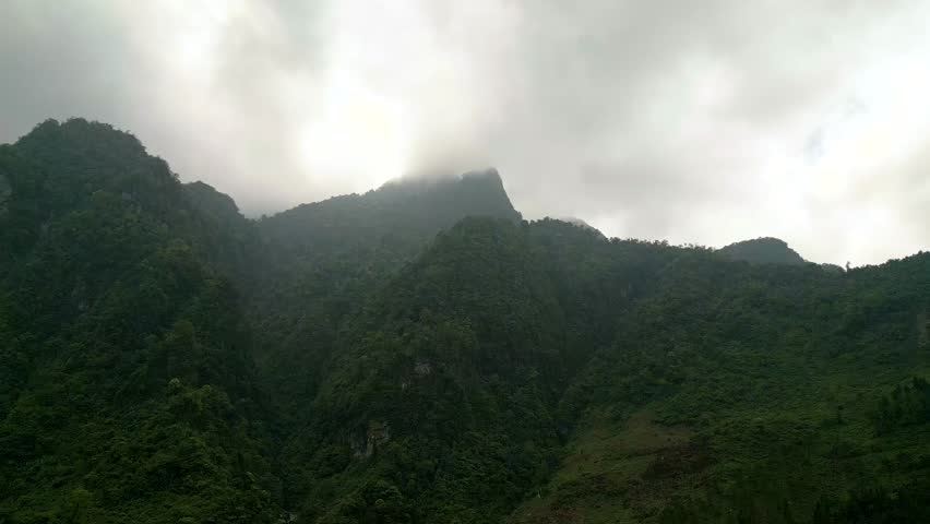 
Drone ascends with a gentle turn over a lush green mountain in Ha Giang, Vietnam. In the background, a tall peak rises into the clouds as bright sunlight breaks through the mist.