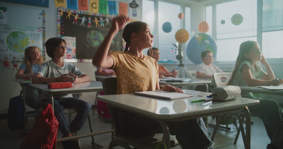Elementary School Students Sitting at the Desks, Listening to Lecture from Teacher, Raising Hands to Provide Correct Answer. Team of Smart Diverse Kids Studying Science, Geography in Modern Classroom.