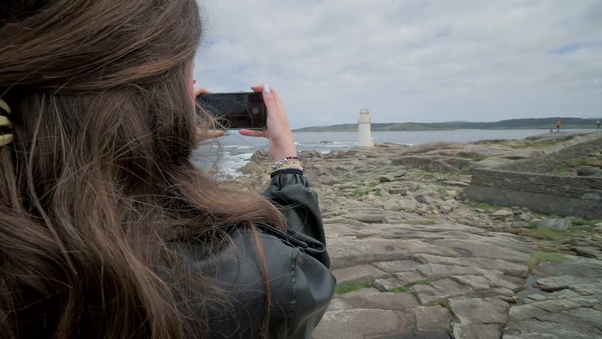 Female photographer capturing scenic coastal landscape of Muxia lighthouse, standing on rocky shoreline with cloudy sky overlooking Atlantic ocean in Galicia, Spain