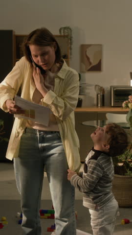 Vertical shot of little son distracting mother from work pulling her shirt and yelling, she talking on phone holding business papers and asking him for silence at home