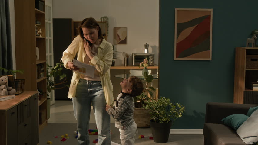 Naughty boy pulling mothers shirt to seek for attention when she speaking on phone holding documents during remote workday