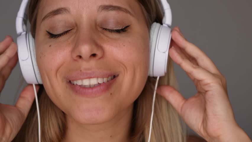 Close-up of a young blonde woman wearing headphones listening to music, dancing, singing and enjoying. She moves in rhythm with closed eyes and a blissful smile. Pure joy and musical flow