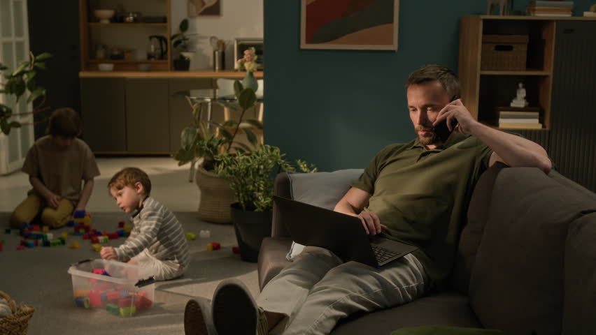 Young man with laptop on his lap having phone conversation when sitting on couch and working remotely, two boys playing on floor in living room