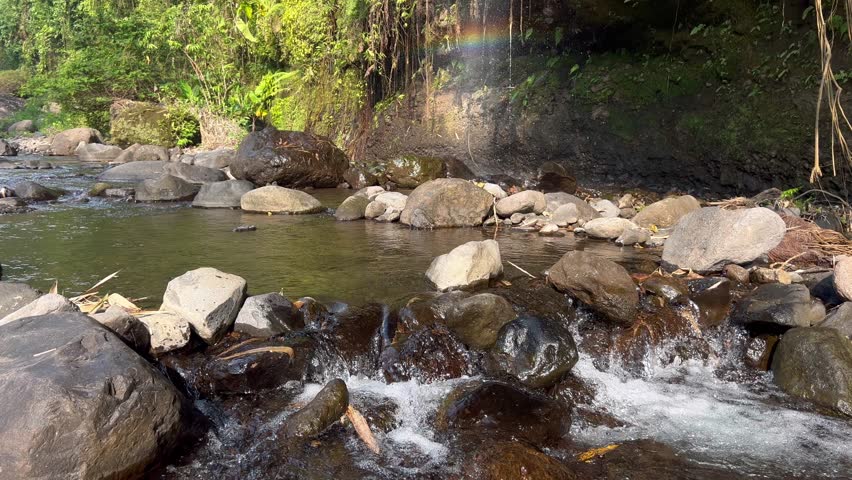The foam of the river's water, from the calm current, resembles pearls. The backdrop is a curtain waterfall, reflected by sunlight, creating a dancing rainbow. 4K 24 FPS.