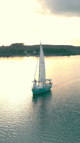 Aerial shot of a single yacht sailing at sunset across open sea. The golden sun reflects off calm water as the drone captures the sailboat from above.
