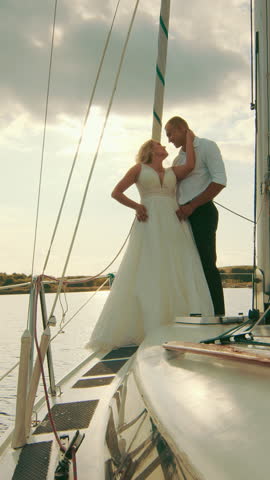 Couple stands on yacht bow during sunset. They embrace while the golden sky glows behind them, repeating the famous Titanic scene in a romantic moment.