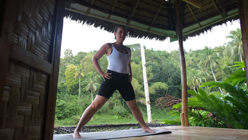 Engaging in yoga poses on a bamboo hut terrace, the young woman embraces the calmness of nature. Vibrant greenery create a perfect backdrop for wellness, slow motion