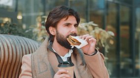 Indian young businessman tourist enjoy lunch break eating tasty sandwich meal and drinking hot beverage coffee or tea. Arabian bearded man guy relaxing sitting on a bench on downtown city street - Powered by Shutterstock - Get 15% off with code: PIKWIZARD15