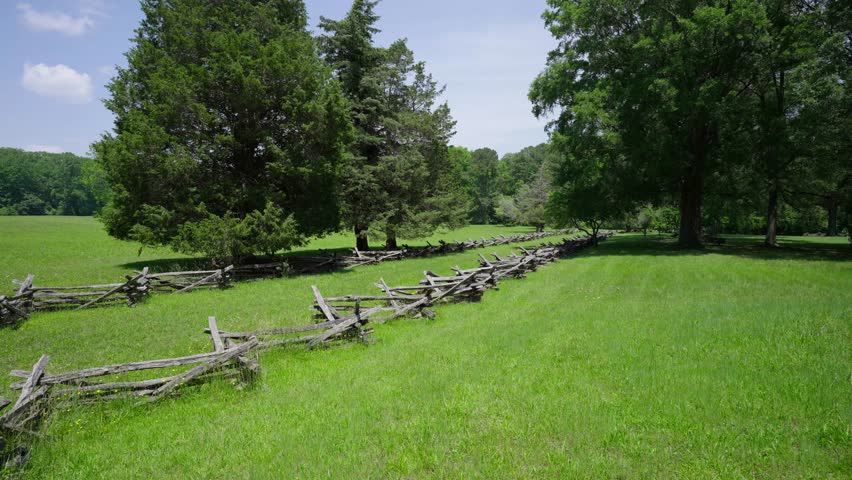 Historic split rail fencing lining a grassy Yorktown battlefield with trees and open fields under a bright summer sky in Yorktown, Virginia.