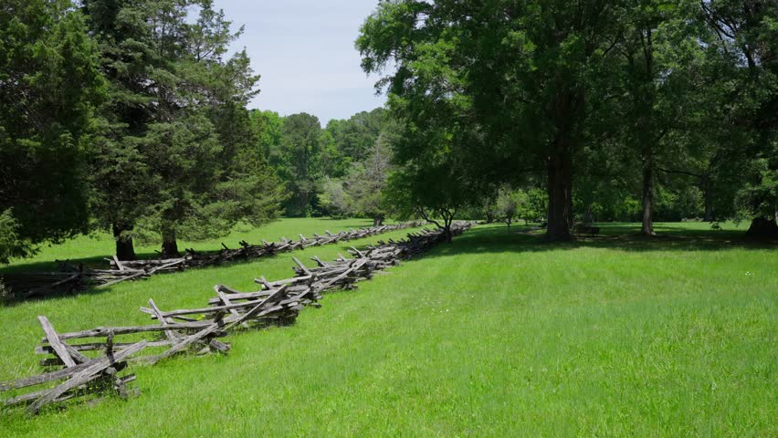 Historic split rail fencing lining a grassy Yorktown battlefield with trees and open fields under a bright summer sky in Yorktown, Virginia.