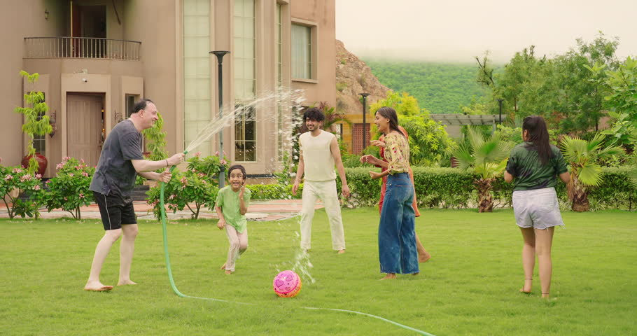 Indian happy father hold hose pipe spraying water on extended family group playing games in outdoor house backyard lawn having fun joy spend funny time enjoying summer vacation together outside park