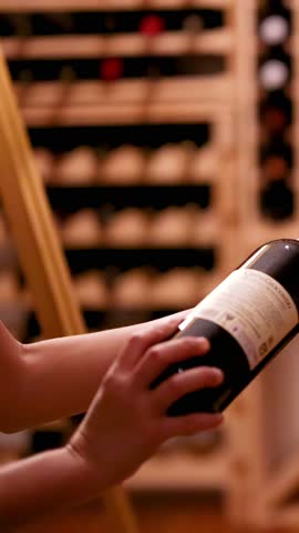 A person carefully inspects a red wine bottle in a warmly lit wine cellar, rotating it to read the label. Shallow depth of field, steady camera